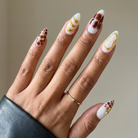 Hand with intricately designed nails featuring white, gold, and brown patterns against a neutral background.